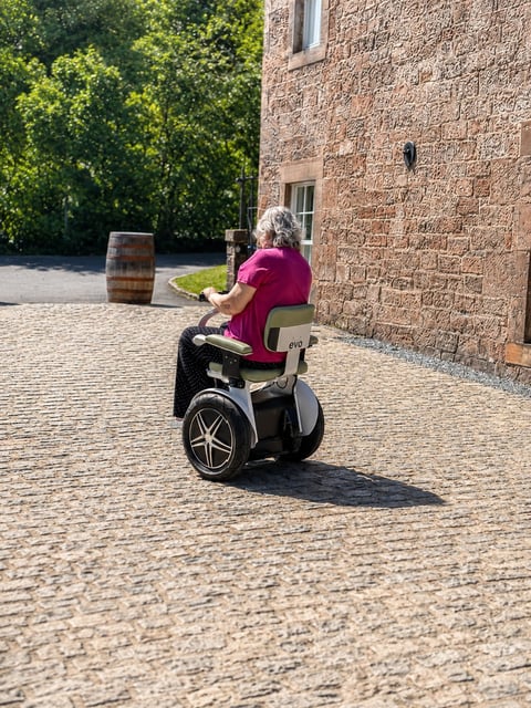Elderly woman in magenta shirt riding a Segway on a brick courtyard next to a stone building